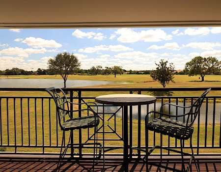A sunny balcony view overlooks a grassy field with scattered trees, a round table and two chairs framed by a black railing.