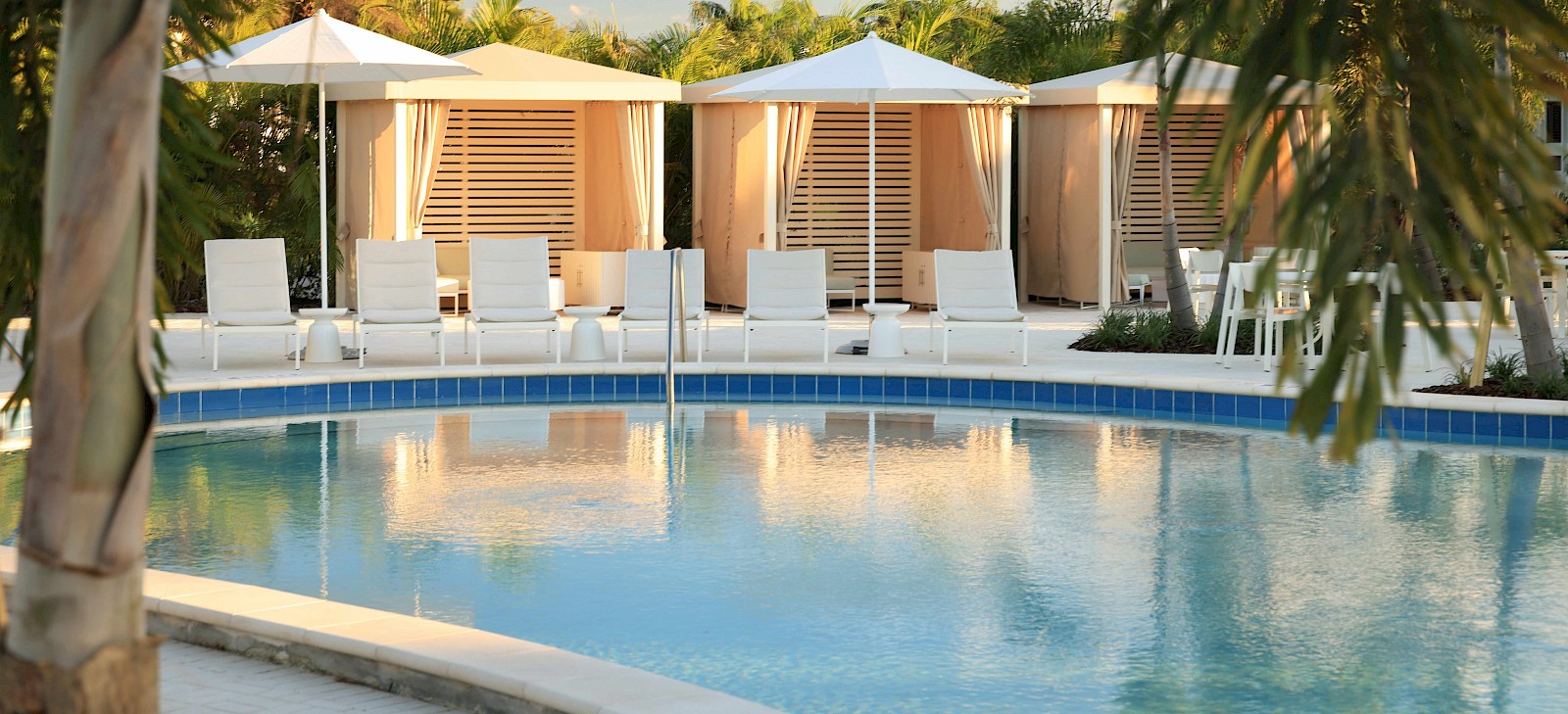 Relaxing poolside scene with white lounge chairs and cabanas surrounded by greenery.