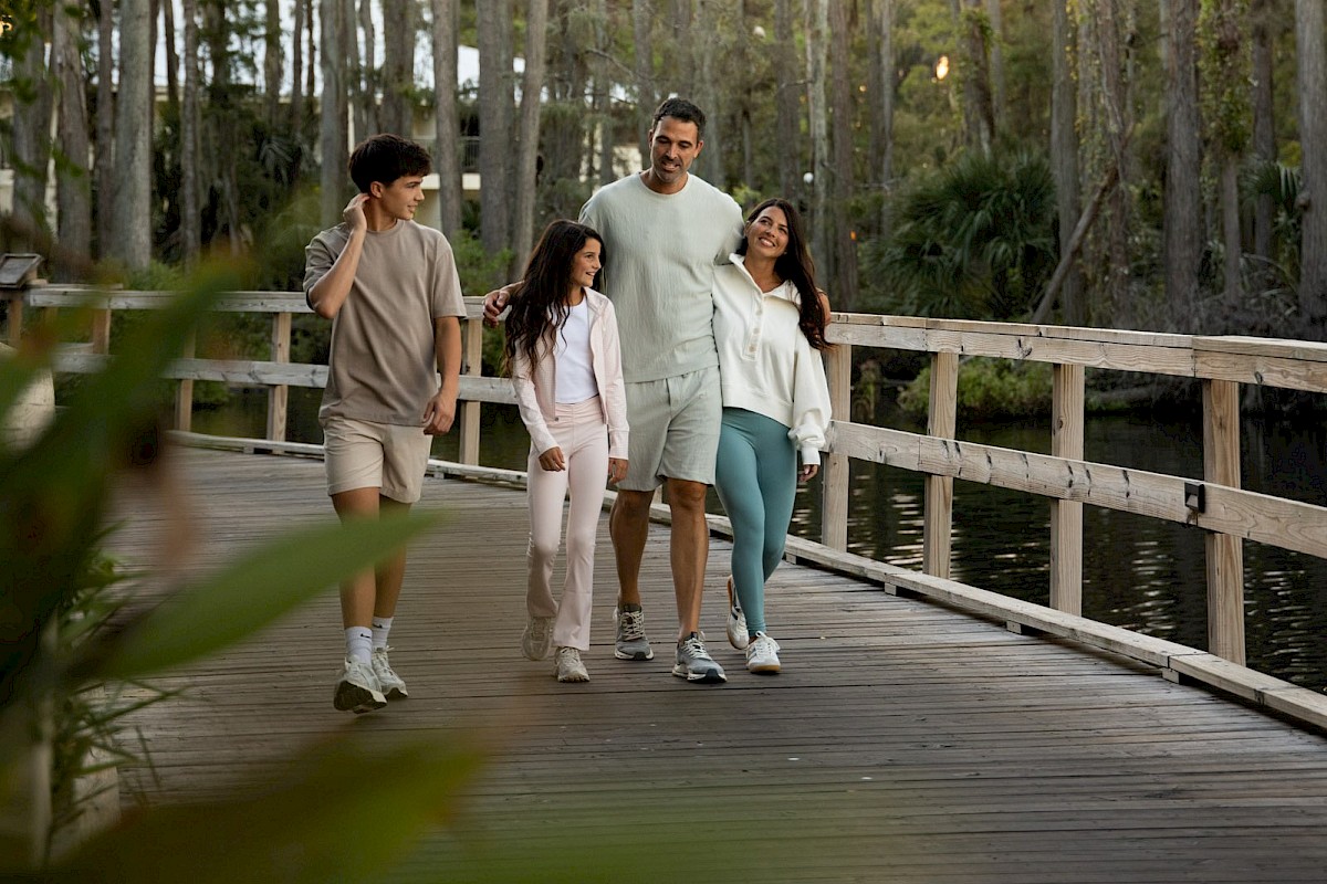 A family of four walks happily along a wooden bridge in a lush, green forest during sunset.