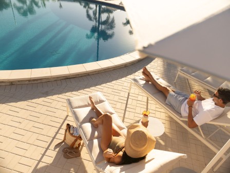 A couple lounges by a pool on sunbeds, with drinks on a table. The scene is sunny and relaxing.