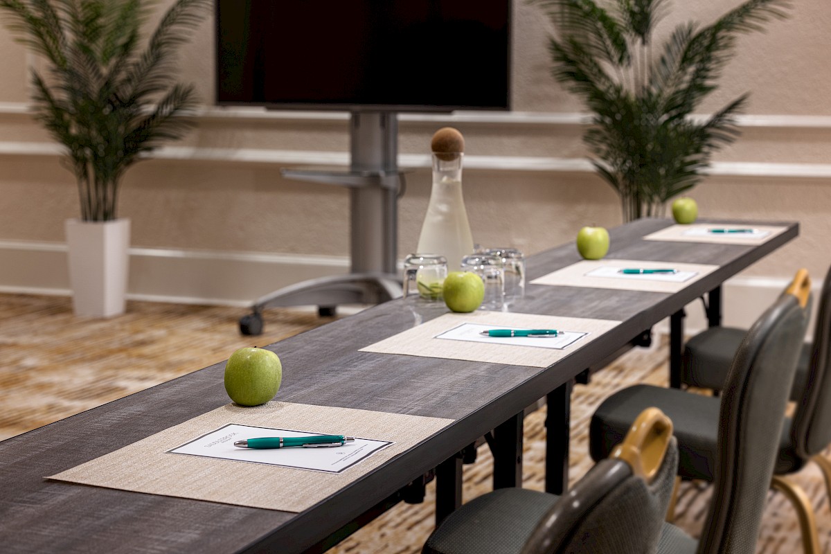 This image shows a conference table set with green apples, pens, paper, glasses, a water bottle, and a carafe in a meeting room.