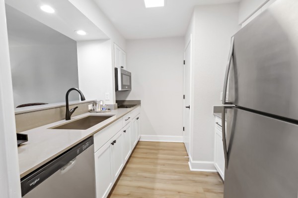 A modern, minimalistic kitchen with white cabinets, stainless steel appliances, light wood flooring, and recessed lighting.