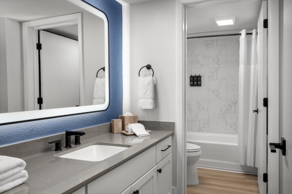 A modern bathroom featuring a vanity with a mirror, towels, and a soap dispenser, adjacent to a shower with a curtain and a toilet.