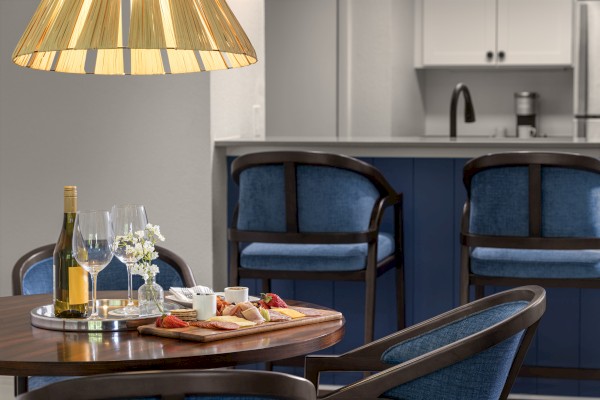 A cozy dining area shows a wooden table with snacks, wine, and glasses next to blue upholstered chairs and a modern kitchen in the background.