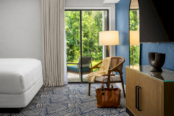 A cozy hotel room with a white bed, a sliding glass door to a green outdoor space, and stylish furniture including a chair, lamp, and TV.