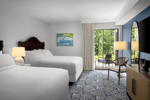 A modern hotel room with two beds, a sitting area outside, and vibrant greenery visible through a sliding glass door.