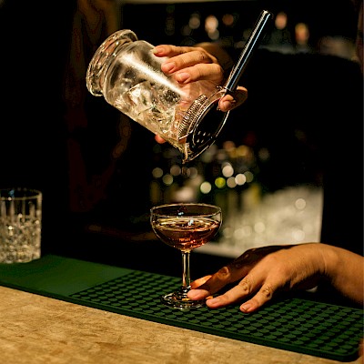 A bartender is pouring a cocktail from a shaker into a coupe glass on a bar counter.