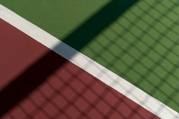 A shadow of a fence falls over a red and green tennis court with a white line dividing the two sections.