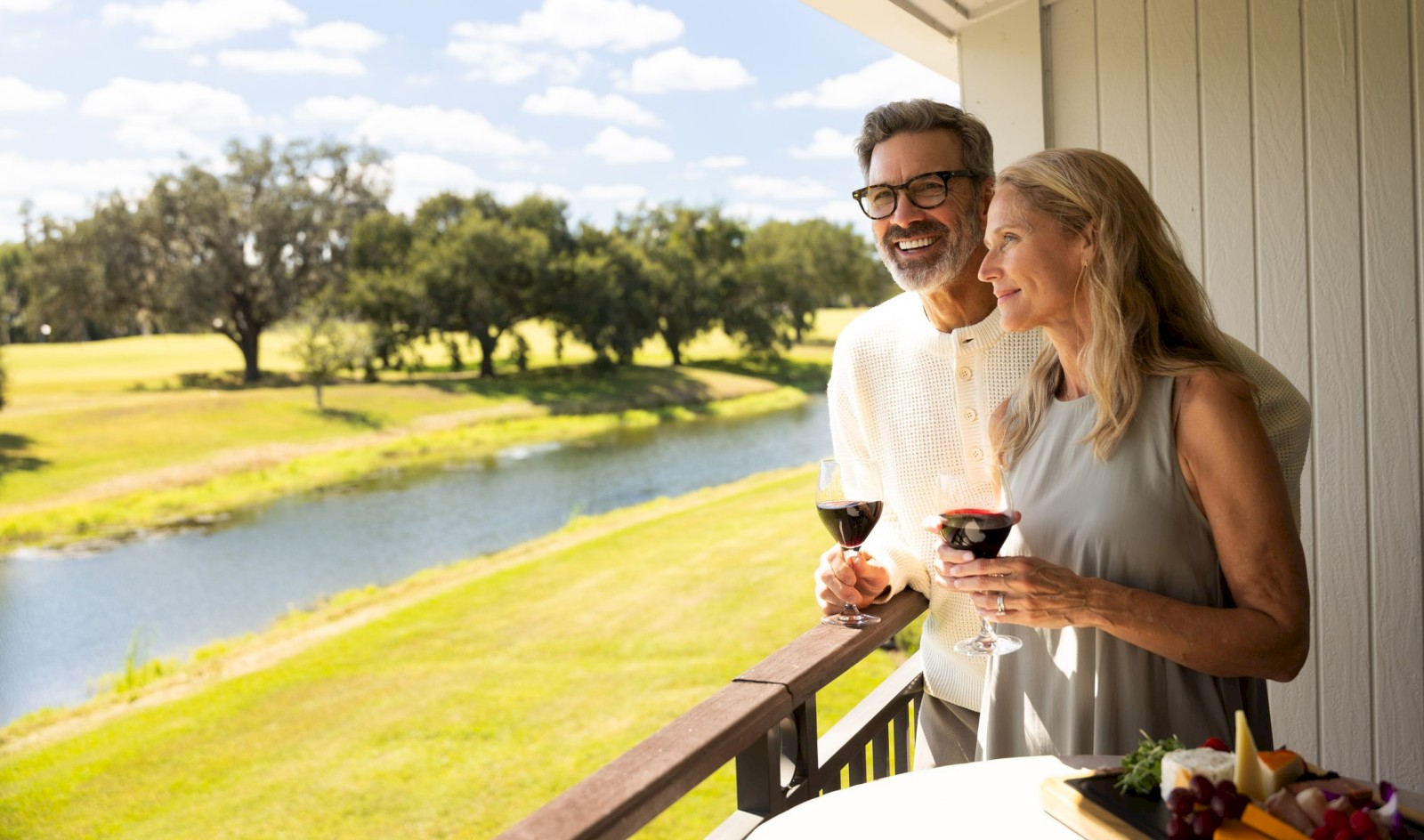 A couple enjoys a scenic view from a balcony with wine glasses in hand, overlooking a lush green landscape and a small river.