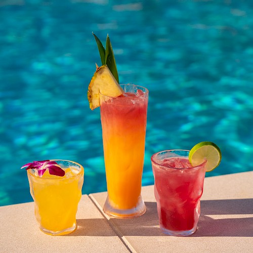 Colorful tropical cocktails with fruit garnishes are set beside a swimming pool, creating a perfect summer poolside scene.
