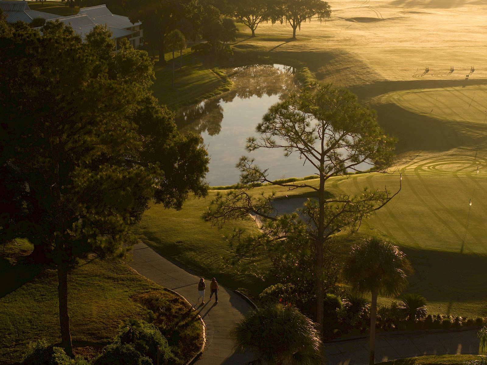 A peaceful golf course scene at sunrise with a pond, trees, and pathways, observed from an elevated perspective.