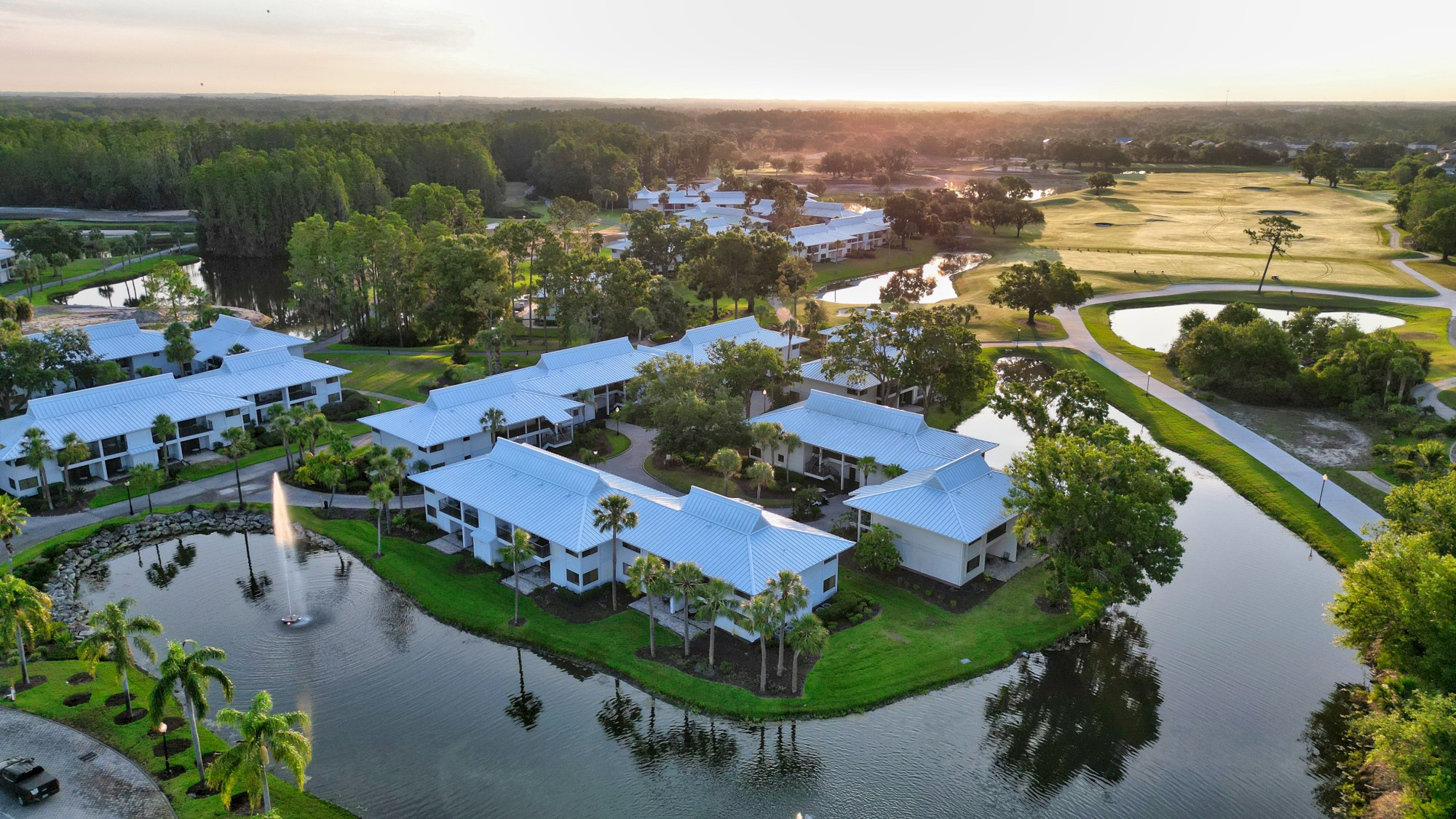 An aerial view of a serene residential area with white-roofed buildings, surrounded by water and greenery, under a sunset sky.