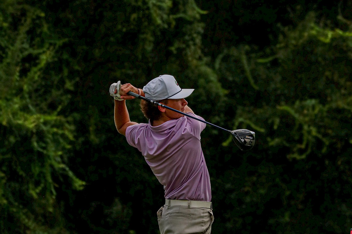 A person is swinging a golf club on a golf course, wearing a cap, shorts, and a polo shirt. The background is lush with greenery.