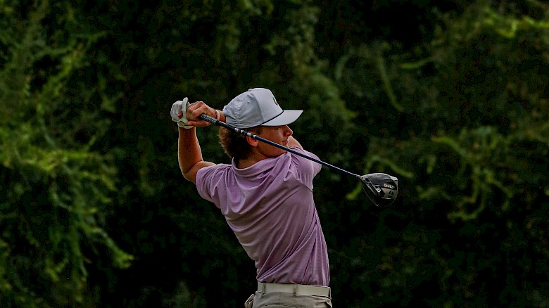 A person is swinging a golf club on a golf course, wearing a cap, shorts, and a polo shirt. The background is lush with greenery.