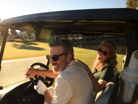 A smiling older man and woman enjoying a sunny day riding a golf cart by a pond.