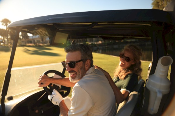 A smiling older man and woman enjoying a sunny day riding a golf cart by a pond.