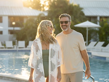 A couple walks happily by a pool on a sunny day, dressed in summer attire, enjoying a relaxing moment together.