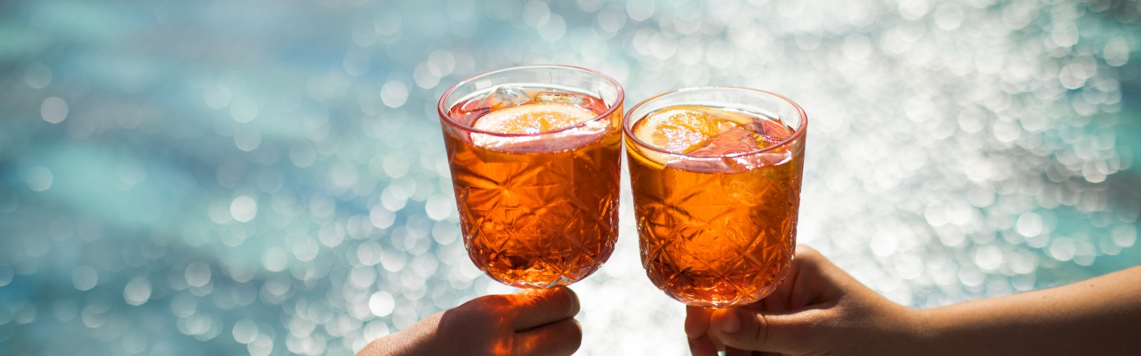 Two hands holding glasses with orange drinks, clinking them together in front of a sparkling pool background.