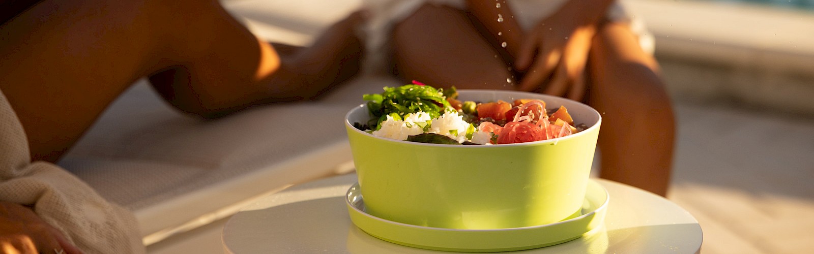 A person squeezes lemon over a bowl of salad on a table, with poolside vibes and relaxed people in the background.