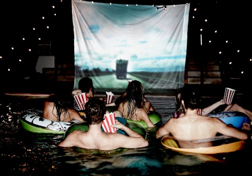People are floating in a pool watching a movie on a large outdoor screen at night.