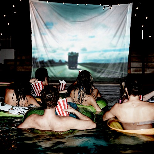 People are floating in a pool watching a movie on a large outdoor screen at night.