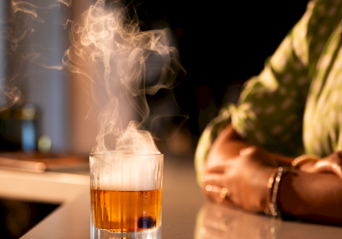 A glowing drink with smoke rises from a clear glass on a reflective surface, with a person wearing a polka dot shirt nearby.