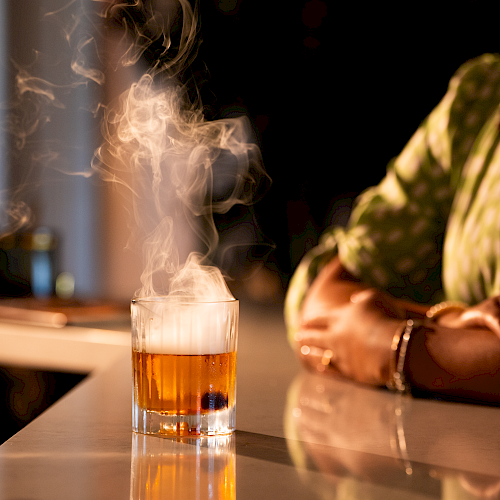 A glowing drink with smoke rises from a clear glass on a reflective surface, with a person wearing a polka dot shirt nearby.