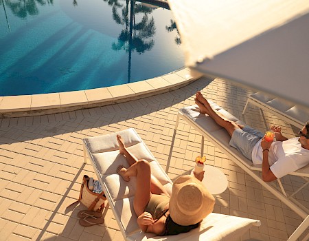 A couple relaxes by a pool with tropical drinks, lounging on chairs under an umbrella on a sunny day.