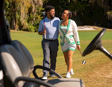 Two people smiling and walking on a golf course, holding golf clubs and enjoying each other's company.
