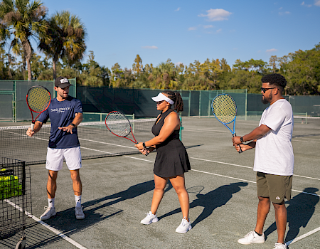 People are on a tennis court during a lesson, holding rackets, with tennis balls in a basket nearby, under a clear, sunny sky.