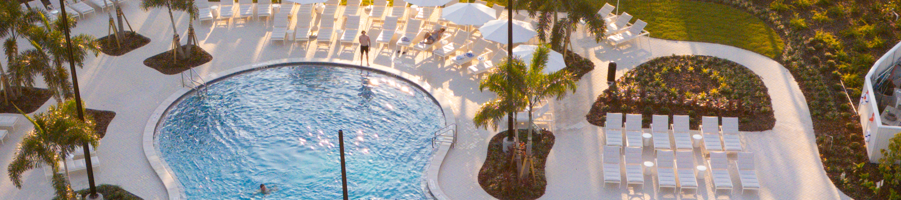 Aerial view of a hotel pool area with loungers, umbrellas, palm trees, and people enjoying the pool in a tropical setting.
