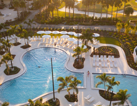 Aerial view of a hotel pool area with loungers, umbrellas, palm trees, and people enjoying the pool in a tropical setting.