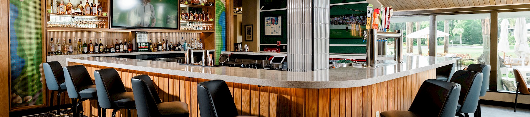 A modern bar with a wooden counter, black chairs, a TV, and outdoor seating visible through large windows, creating a cozy yet stylish atmosphere.