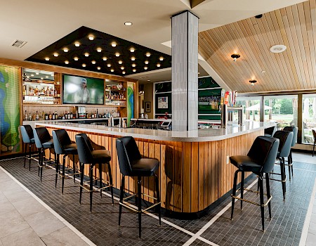 A modern bar with a wooden counter, black chairs, a TV, and outdoor seating visible through large windows, creating a cozy yet stylish atmosphere.