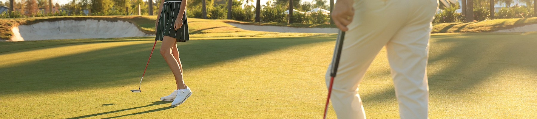 A man and woman are playing golf on a sunny course with palm trees, the woman appears to be close to sinking a putt while the man observes.