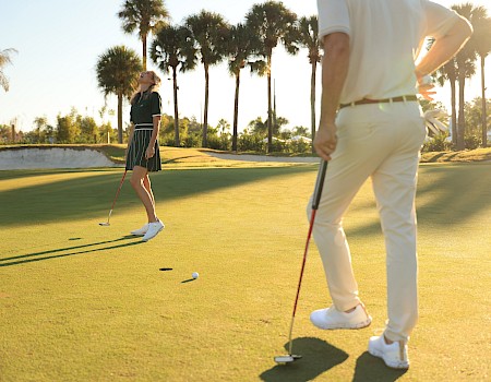 A man and woman are playing golf on a sunny course with palm trees, the woman appears to be close to sinking a putt while the man observes.