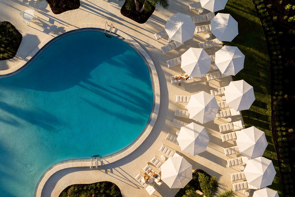 Aerial view of a poolside with sun loungers, white umbrellas, and lush greenery.