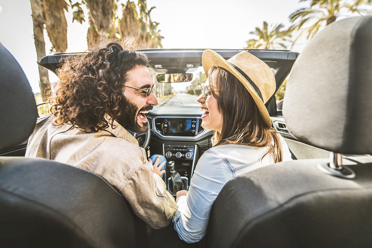 A smiling couple enjoys a fun moment together in a convertible car on a sunny day, surrounded by palm trees and a clear sky.