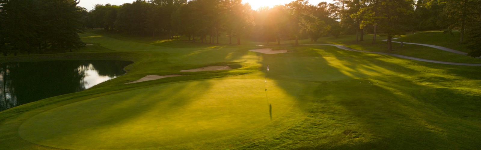 A golf course at sunset, with lush green fairways, sand traps, and a water hazard, surrounded by trees.