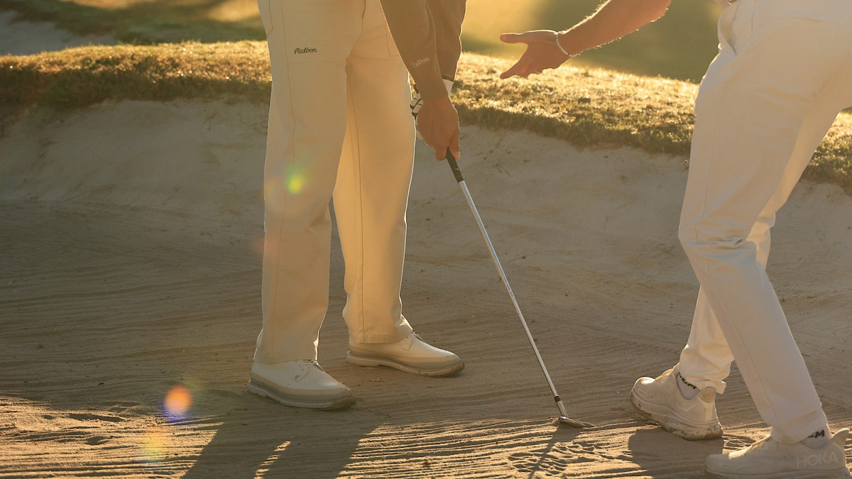 Two golfers on a sunny course, lining up a putt near palm trees.