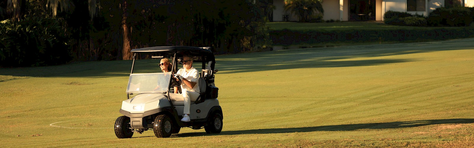 Two people ride a small golf cart across a sunny golf course, with a building and trees in the background.