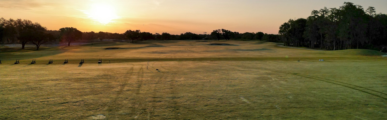 A golf course during sunrise with an open field, trees on the sides, and a few golf carts in the distance.