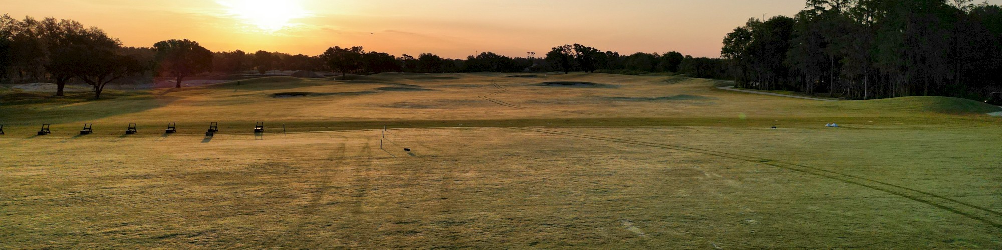A golf course during sunrise with an open field, trees on the sides, and a few golf carts in the distance.