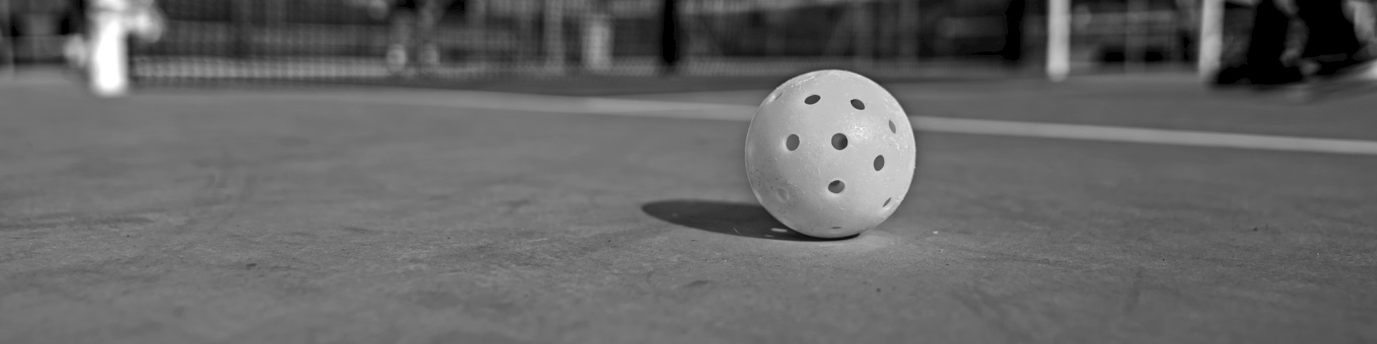 A pickleball sits on a court with a net and players in the background, captured in black and white.