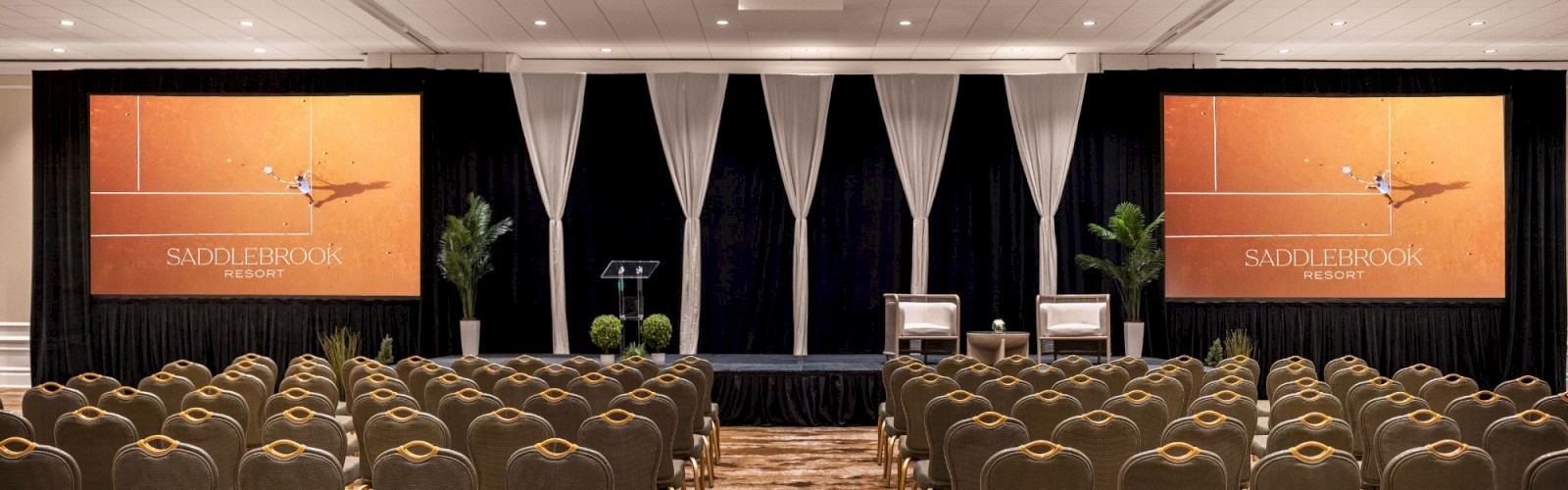 An empty conference room is set up with rows of chairs facing a stage with two screens labeled 