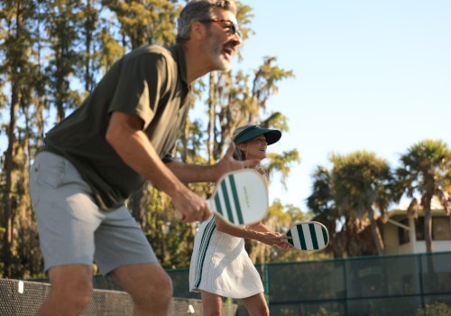 Two seniors play pickleball outdoors on a sunny day, smiling and holding paddles while enjoying the game.