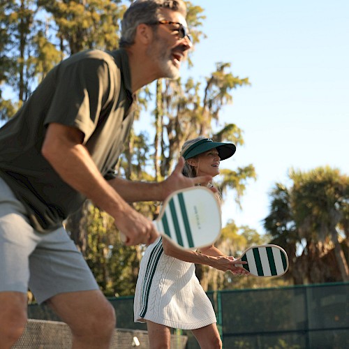 Two seniors play pickleball outdoors on a sunny day, smiling and holding paddles while enjoying the game.