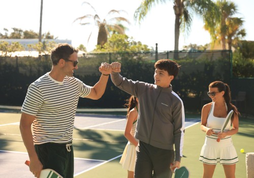 Two people fist bump on a tennis court, surrounded by women holding rackets, during a sunny outdoor tennis game.