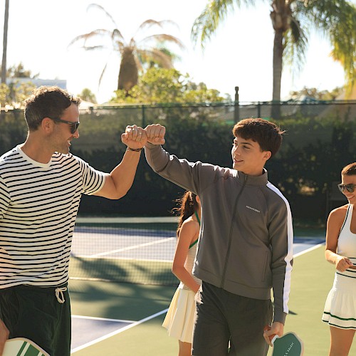 Two people fist bump on a tennis court, surrounded by women holding rackets, during a sunny outdoor tennis game.