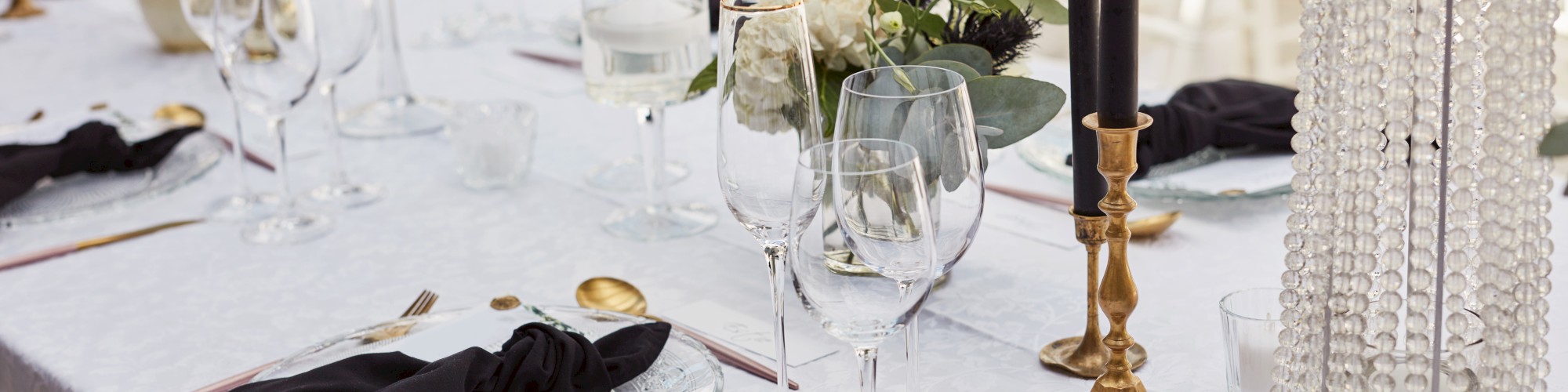 A formal table setting with glassware, black napkins, gold cutlery, candles, and floral centerpiece on a white tablecloth.